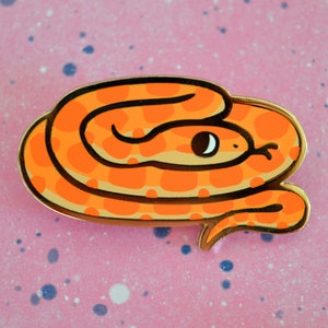 May include: An orange snake enamel pin with a gold border. The snake has a light orange belly and darker orange spots. The pin is set against a pink background with blue and white speckles. The snake has a black eye and a white highlight.