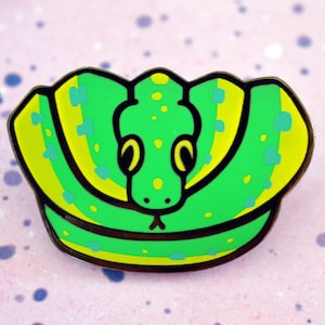 May include: Green and yellow enamel pin of a cartoon snake curled up with its head facing forward. The snake has black eyes and a yellow tongue.