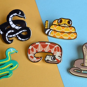 Op de afbeelding: Vijf emaillepins met verschillende soorten slangen. De pins zijn kleurrijk en gedetailleerd, elke slang heeft een uniek ontwerp. De slangen zijn een zwart-witte slang, een groene slang, een rood-witte slang, een geel-oranje slang en een bruine slang.