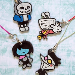 Peut inclure: Quatre breloques de personnages de dessins animés avec des fermoirs argentés et des cordons colorés. Les breloques comprennent un squelette en veste bleue, une tasse blanche avec un design rouge et jaune, un personnage aux cheveux noirs et à la peau bleue et un personnage au visage blanc et aux cheveux noirs.