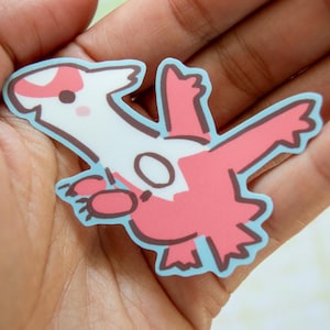 May include: A die-cut sticker of a cartoon character with a white body, pink wings, and a light blue outline. The character has a small, round face with pink cheeks and a brown outline. The sticker is held in a hand.