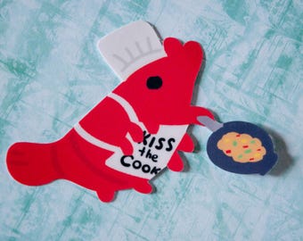 May include: A red lobster wearing a white chef's hat and apron holding a blue pan with scrambled eggs. The lobster is holding a spatula and has the text "Kiss the Cook" on its apron.