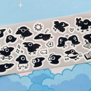May include: A sheet of black and white crow stickers with various designs. The stickers feature crows in different poses, including sleeping, reading, skateboarding, and wearing sunglasses. Some stickers also include small details like stars, flowers, and musical notes.