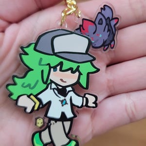 Op de afbeelding: Een heldere acryl bedel met een stripfiguur met groen haar, een witte jas en een grijze hoed. Een klein paars wezen met rode accenten is bevestigd. De bedel wordt vastgehouden door een goudkleurige ketting.