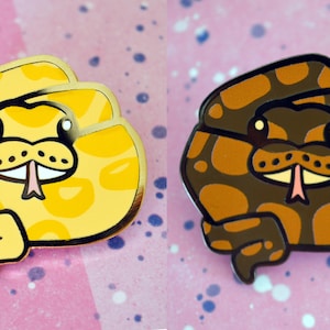 Op de afbeelding: Twee emaillepins met cartoon-slangen. De slang links is geel met zwarte vlekken en de slang rechts is bruin met zwarte vlekken. Beide slangen hebben zwarte ogen, een roze tong en witte tanden.