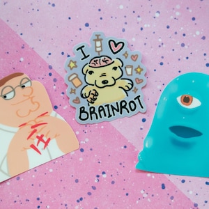 May include: A collection of stickers featuring cartoon characters and designs. One sticker depicts a character with the text "I ❤️ BRAINROT". Another shows a blue, one-eyed creature. The third sticker is a cartoon character with Japanese text.