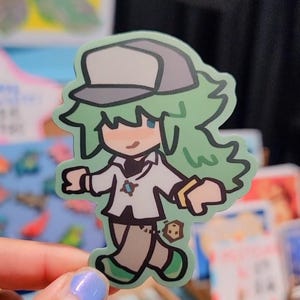 Op de afbeelding: Een sticker met een stripfiguur met groen haar, een grijze pet en een wit overhemd. Het personage heeft een kleine glimlach en is zwart omlijnd. De sticker wordt vastgehouden door een persoon met lichtblauwe nagellak.