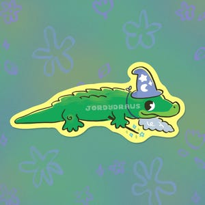 Może przedstawiać: Zielony aligator w niebieskim kapeluszu czarodzieja z gwiazdami i rogalem księżyca. Aligator trzyma magiczną różdżkę z gwiazdami i chmurą na końcu. Tekst "JORDYDRAWS" jest napisany na ciele aligatora.
