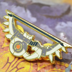May include: A gold-coloured metal pin in the shape of a bow and arrow. The pin features a grey bow with white accents and a gold arrow with a black stripe. The pin has a decorative design with leaves.