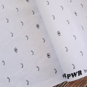 Può includere: Tessuto bianco con un motivo ripetitivo di occhi e ciglia neri. Il tessuto presenta il testo "PWR" in lettere nere.