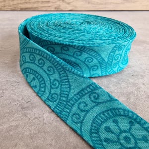 Op de afbeelding: Een rol turquoise stoffen lint met een herhalend cirkelvormig patroon. Het lint is afgerold en toont het gedetailleerde ontwerp. De stof heeft een getextureerde uitstraling en de kleur is een levendig teal.