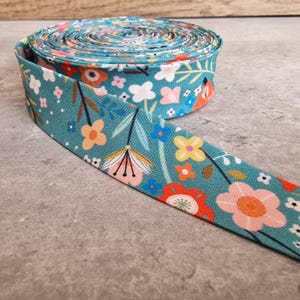 May include: A roll of floral fabric ribbon in teal, pink, orange, yellow, and white. The ribbon features a repeating pattern of flowers and leaves. The ribbon is unrolled, showing the pattern. The fabric is likely cotton.