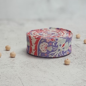 Pode incluir: Um rolo de fita de tecido com padrão floral roxo, rosa e vermelho. A fita está enrolada e colocada sobre uma superfície branca com contas de madeira espalhadas ao redor.