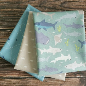 May include: Three fabric panels featuring different shark designs. The top panel is light blue with a white dashed line pattern and small white sharks. The middle panel is beige with a white shark pattern. The bottom panel is light blue with a variety of white, grey, and blue sharks, rays, and hammerhead sharks.