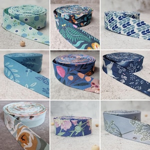 Può includere: Una collezione di nove rotoli di nastro sbieco in tessuto con vari motivi, inclusi disegni floreali, botanici e geometrici in tonalità di blu, verde e bianco. Ogni rotolo è avvolto e mostra un distinto motivo tessile stampato.