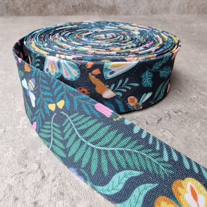 Peut inclure: Un rouleau de ruban en tissu bleu foncé avec un motif floral représentant des papillons, des abeilles et des feuilles. Le ruban est enroulé et présente un motif floral vert, jaune, orange, rose et blanc.