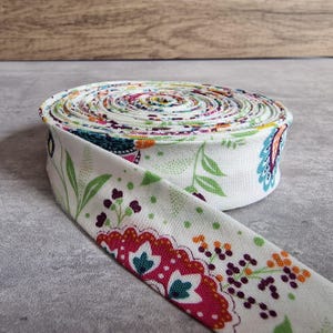 May include: A roll of white fabric bias tape with a colorful floral pattern. The tape features a repeating design of flowers and leaves in shades of pink, green, blue, and orange. The tape is rolled up on a gray surface.