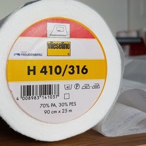 Op de afbeelding: Een witte rol Vlieseline H 410/316 vlieseline met een geel label. Het label heeft een barcode, wasinstructies en de tekst "70% PA, 30% PES 90 cm x 25 m".