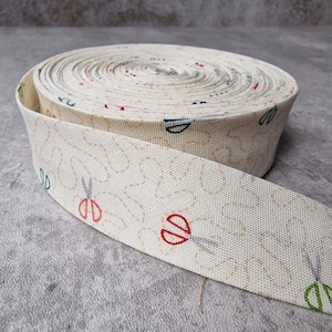 Puede incluir: Un rollo de cinta de tela de color blanco roto con un patrón repetitivo de tijeras y hojas. La cinta está enrollada y el patrón es visible en el exterior del rollo.