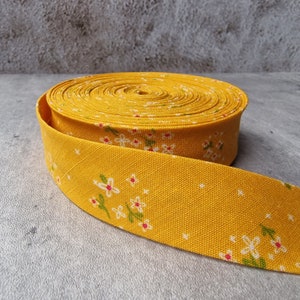 Peut inclure: Un rouleau de ruban biais jaune moutarde orné d'un motif répétitif de petites fleurs blanches avec des feuilles vertes et de minuscules cœurs rouges. Le ruban en tissu présente une texture tissée visible et est enroulé en bobine circulaire.
