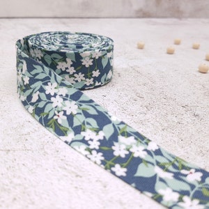 May include: A roll of blue and white floral ribbon with a white background and small white flowers on green stems. The ribbon is rolled up and has a few inches of ribbon unrolled.