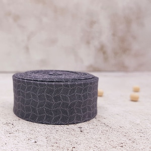 May include: A roll of dark gray fabric with a white geometric pattern. The fabric is rolled up and sitting on a light gray surface.