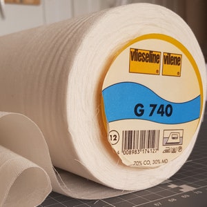 Könnte beinhalten: Eine Rolle weißen Stoffes mit einem gelben Etikett, auf dem "vlieseline vilene" und "G 740" steht. Das Etikett enthält auch einen Barcode und den Text "70% CO, 30% MD".