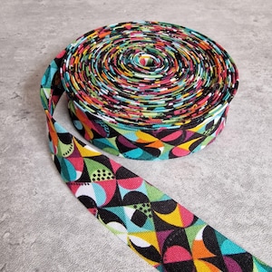 Pode incluir: Um rolo de viés colorido com um padrão geométrico vibrante. A fita apresenta um design repetido de formas pretas, rosa, verdes, amarelas e turquesas. A fita é enrolada firmemente, com uma porção desenrolada, mostrando o padrão.