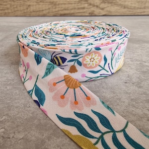 Pode incluir: Fita de tecido enrolada com um padrão floral e de abelhas. O tecido apresenta um fundo creme com flores e folhas rosa, amarelas, azuis e verdes. Uma abelha também é visível no desenho. O tecido está enrolado, com uma parte desenrolada.