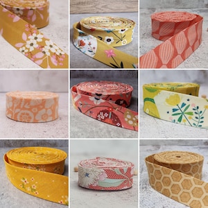 Puede incluir: Una colección de nueve rollos de cinta al bies de tela con varios patrones, incluyendo estampados florales en amarillo, naranja y crema, formas geométricas y un diseño de panal. Cada rollo está enrollado en una bobina circular de tela.