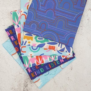 May include: A stack of colorful fabric squares with different patterns and text. The top square is blue with a repeating pattern of arches. The other squares feature a variety of patterns, including flowers, rainbows, and geometric shapes. The text on the squares includes "Free", "Kind", "Listen", and "Brave".