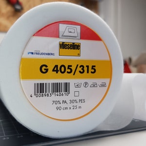 Puede incluir: Un rollo blanco de entretela termoadhesiva Vlieseline G 405/315 para coser. La etiqueta es amarilla con texto negro y un código de barras. Dice "70% PA, 30% PES" y "90 cm x 25 m".