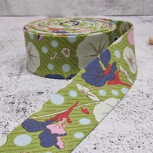 Pode incluir: Um rolo de tecido verde com um padrão floral com flores azuis, rosas e brancas e bolinhas azuis claras. O tecido está enrolado, com uma parte desenrolada, revelando o desenho. O tecido é provavelmente algodão ou similar.