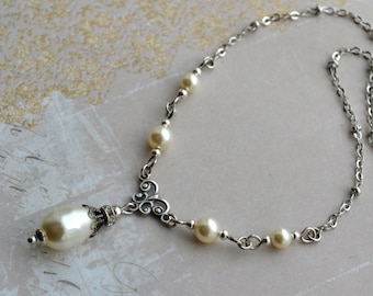 May include: A silver chain necklace with a pendant featuring a decorative design and a large white pearl drop. The necklace is accented with smaller white pearls.