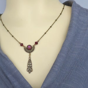 May include: A bronze necklace with a pink crystal pendant. The pendant is shaped like a crescent moon with a teardrop-shaped dangle. The necklace is adorned with small pink crystals.