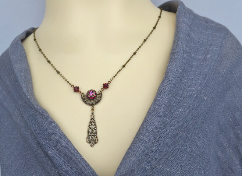 May include: A bronze necklace with a pink crystal pendant. The pendant is shaped like a crescent moon with a teardrop-shaped dangle. The necklace is adorned with small pink crystals.
