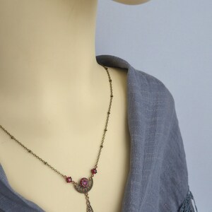 May include: A bronze necklace with a pendant featuring a crescent moon design with a red rhinestone in the center. The pendant is attached to a delicate chain with small bronze beads.