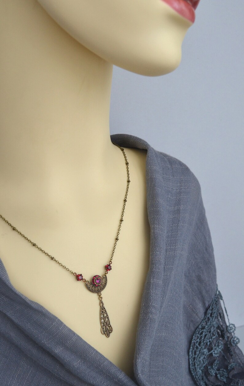 May include: A bronze necklace with a pendant featuring a crescent moon design with a red rhinestone in the center. The pendant is attached to a delicate chain with small bronze beads.
