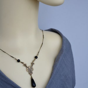 May include: A gold necklace with a black teardrop pendant and black beads. The necklace is on a mannequin wearing a gray shirt.