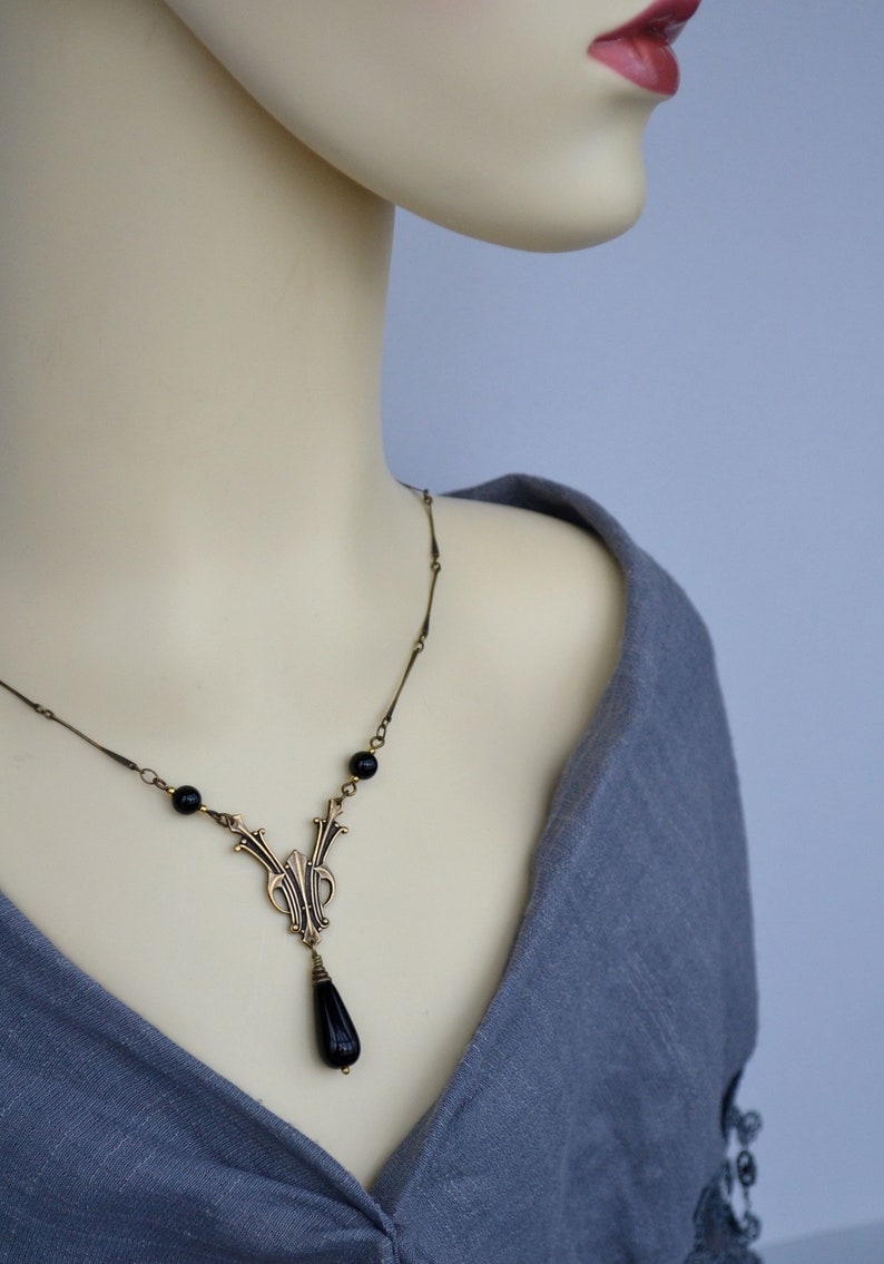 May include: A gold necklace with a black teardrop pendant and black beads. The necklace is on a mannequin wearing a gray shirt.