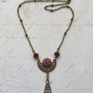 May include: A delicate antique-style necklace with a pendant featuring a crescent moon design with a pink rhinestone center and a teardrop-shaped dangle. The necklace is made of brass and has a chain with small beads.