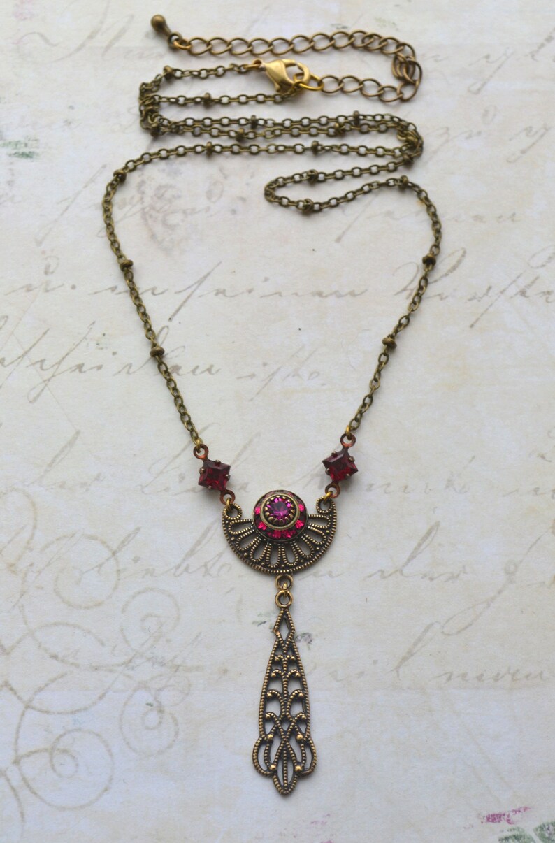 May include: A delicate antique-style necklace with a pendant featuring a crescent moon design with a pink rhinestone center and a teardrop-shaped dangle. The necklace is made of brass and has a chain with small beads.