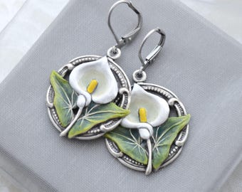 Può includere: Un paio di orecchini rotondi color argento con un design in rilievo a forma di calla. Ogni orecchino presenta un fiore bianco con uno spadice giallo e foglie verdi, inserito in una cornice circolare con chiusura a monachella per la sicurezza.
