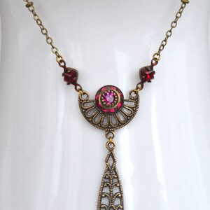 May include: A gold necklace with a pink and red crystal pendant. The pendant is a crescent moon shape with a teardrop hanging from the bottom. The necklace is on a white background.