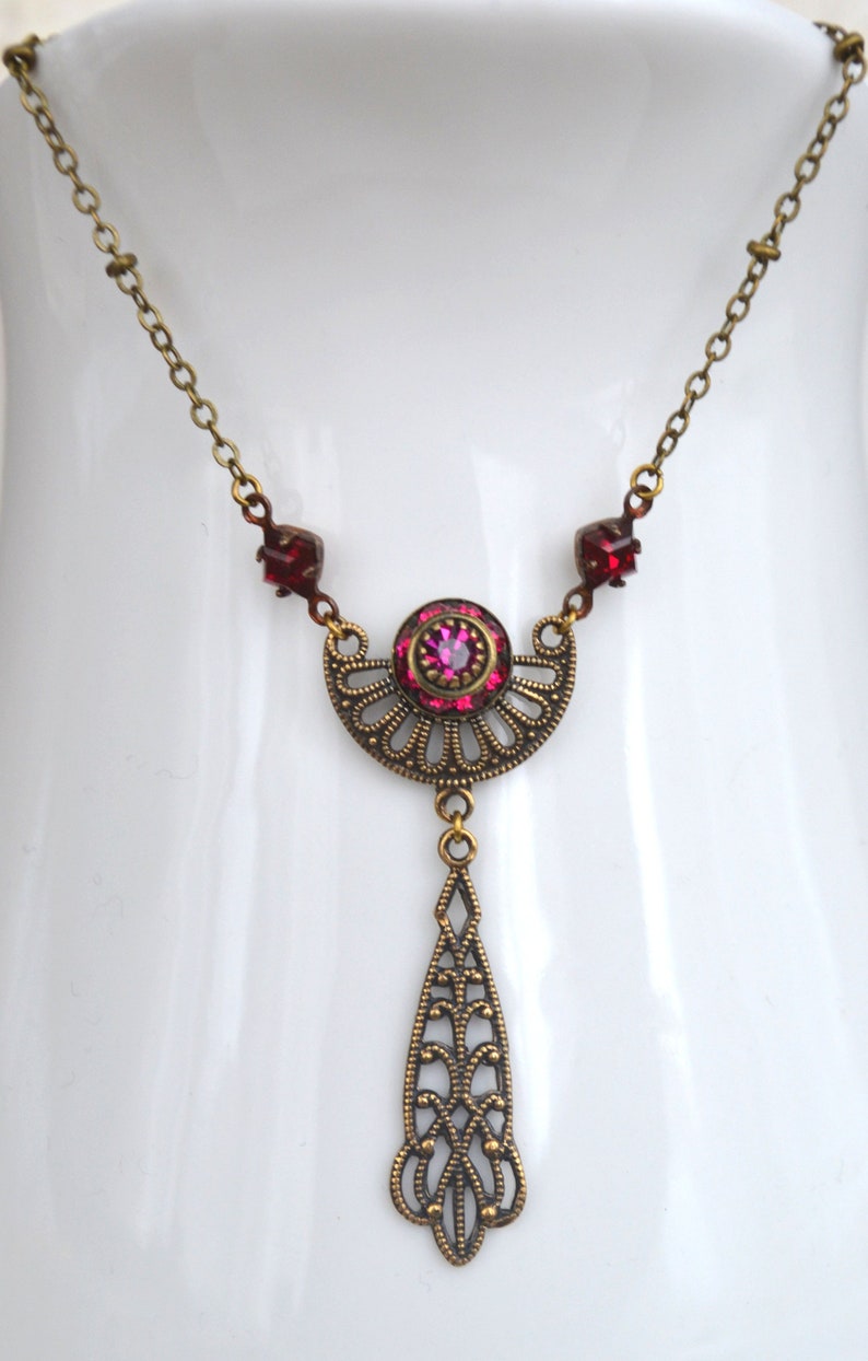 May include: A gold necklace with a pink and red crystal pendant. The pendant is a crescent moon shape with a teardrop hanging from the bottom. The necklace is on a white background.