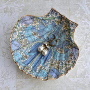 May include: A scallop shell trinket dish featuring a floral pattern in shades of blue, yellow, and green with a gold-painted rim. Resting inside the shell is a pair of drop earrings with bronze-toned hooks and two white teardrop-shaped pearls.