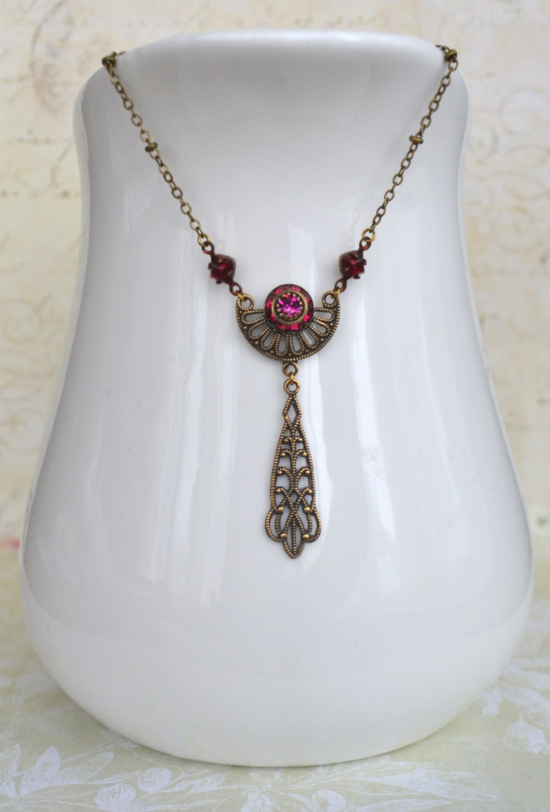 May include: A delicate gold necklace with a pink rhinestone pendant. The pendant features a crescent moon design with intricate filigree details and a dangling drop.