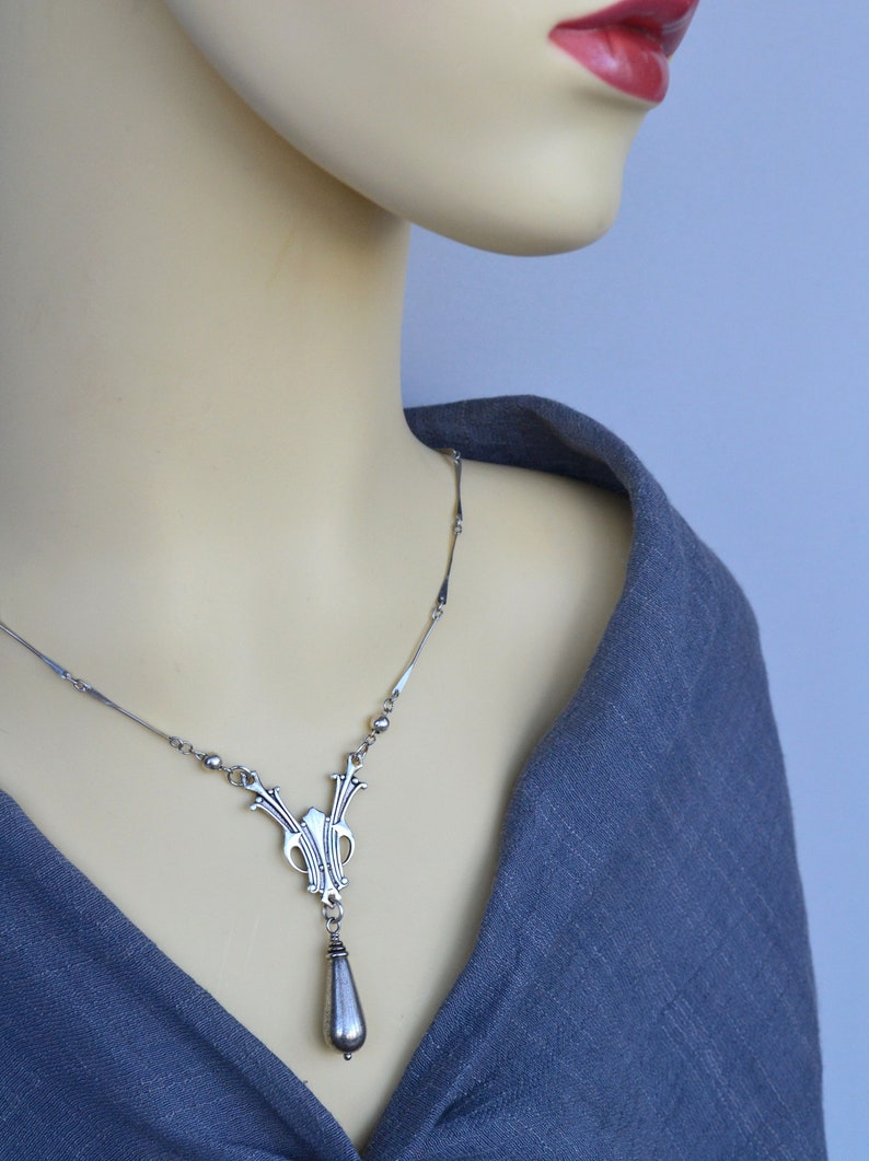 May include: A silver necklace with a teardrop pendant and an intricate design. The necklace is on a mannequin wearing a gray shirt.