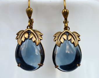 May include: A pair of dangle earrings featuring a teardrop-shaped blue glass stone and gold-toned metal accents. The earrings have a leaf design at the top of the stone.