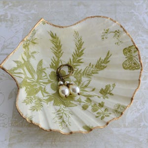 May include: A cream-coloured scallop shell trinket dish featuring a green botanical print with ferns and a butterfly. The edges are trimmed in gold. Resting inside the shell is a pair of drop earrings with bronze-toned hooks and white pearls.